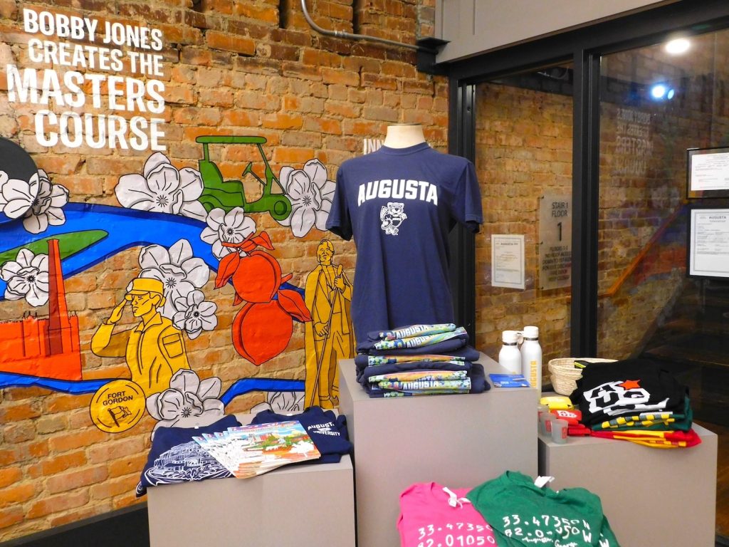 Merchandise inside a small business, including a shirt that says "Augusta" with a college mascot depicting a jaguar on it.