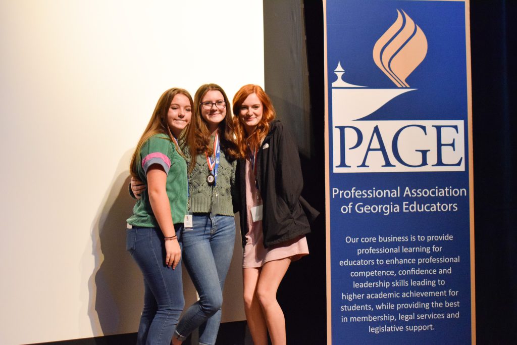 Three female students posing for picture next to a vertical banner with the word "PAGE" in big letters.