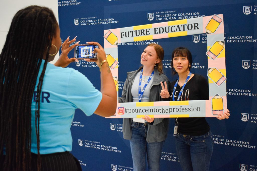 A woman takes a picture of two female students posing while holding an oversized photo frame with pencils and the words "Future Educator."