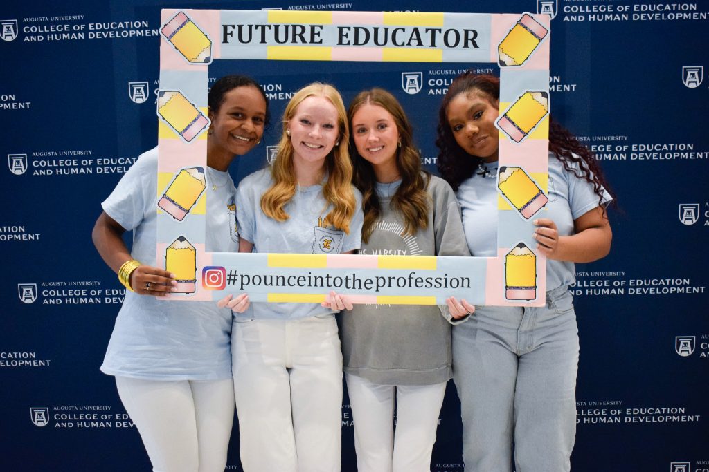 A group of four female students posing for pictures while holding an oversized picture frame with cutouts of pencils around the border and the words "Future Educator."