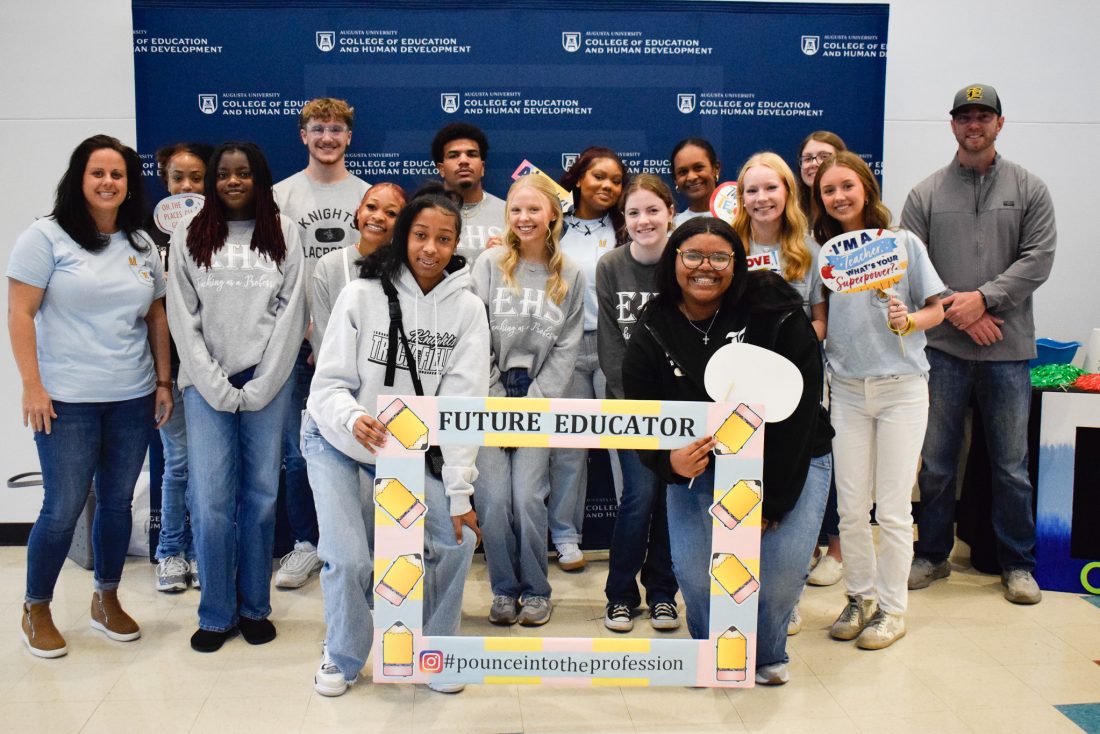 A large group of high school students pose for pictures around an oversized picture frame with cutouts of pencils around the border and the words "Future Educator."