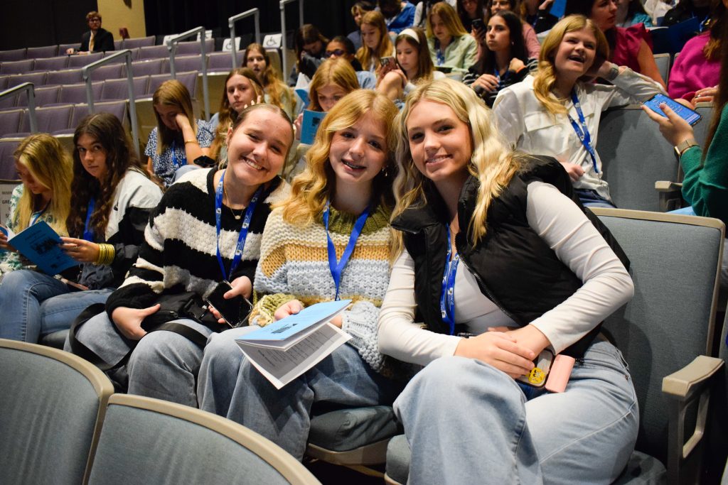 A group of three high school girls in a large auditorium.