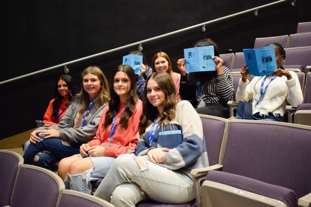 A group of eight high school girls sitting down in a large auditorium.