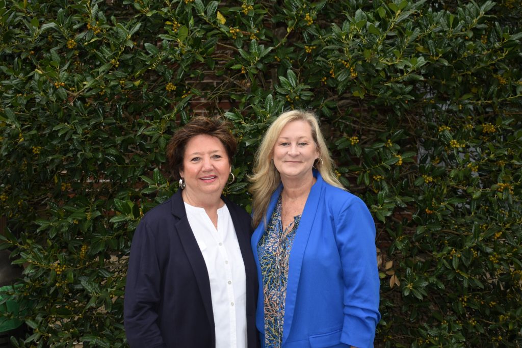 Two women stand side-by-side outside in front of a row of hedges.