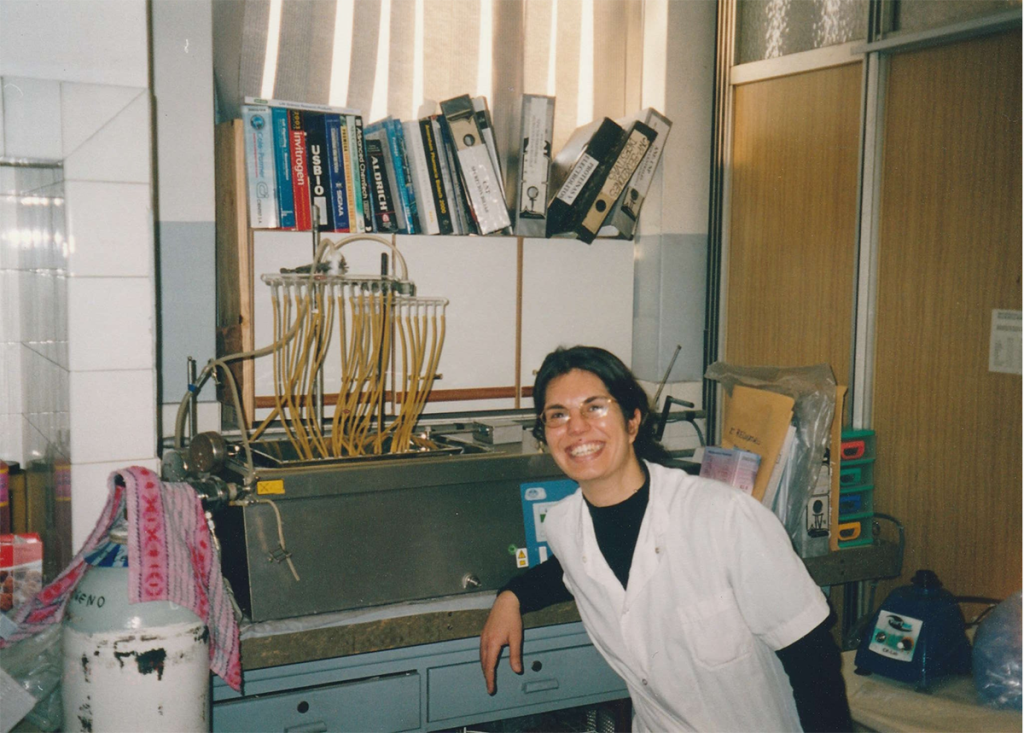 A woman, Dr. Sabbatini, works in an old lab surrounded by equipment with large scientific books on a shelf over her.