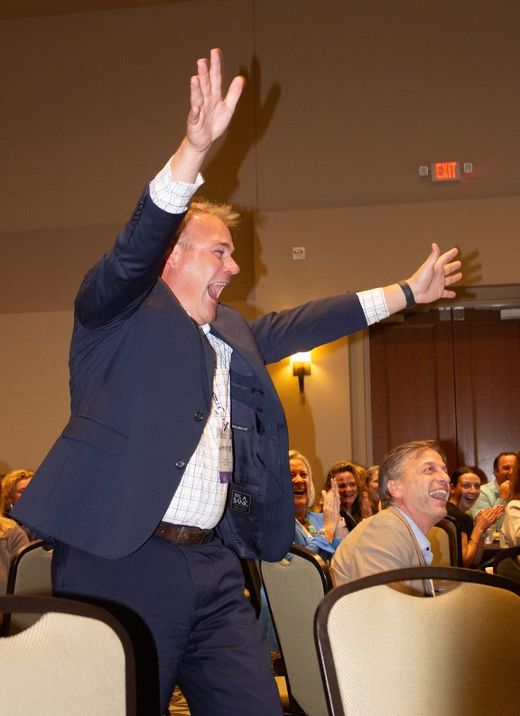 A man holds his hands high as he walks with his mouth wide open in a surprised yell while the audience behind him laughs and claps.