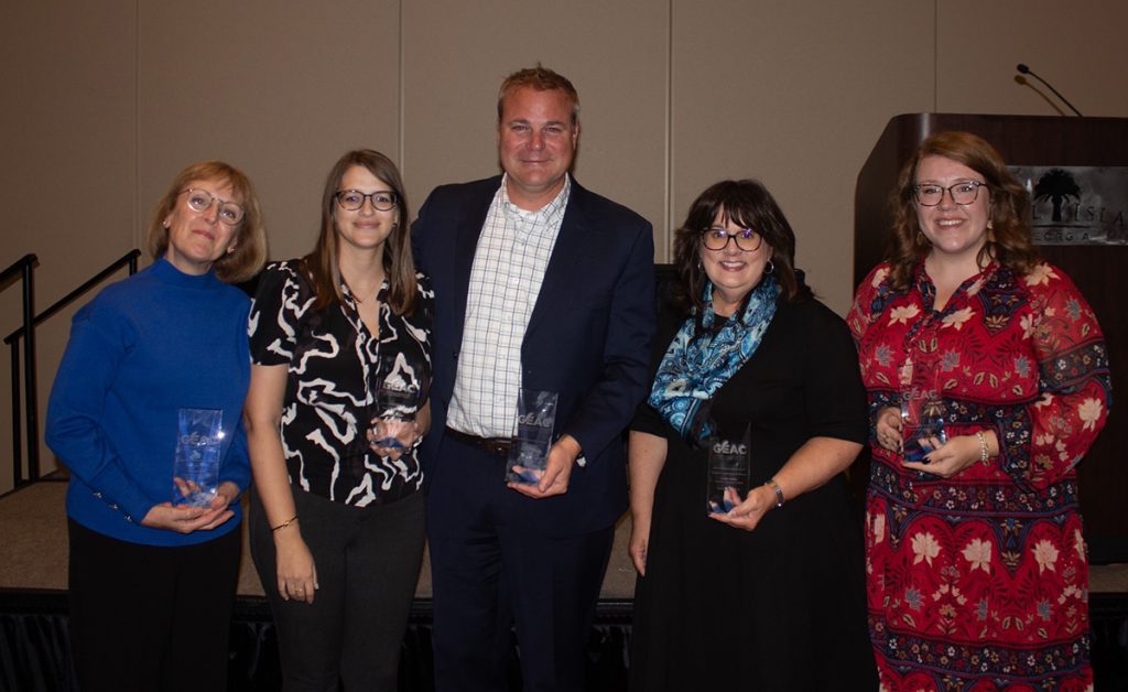 Five people -- four women with a man in the center -- pose near the stage for a photo, holding their awards.