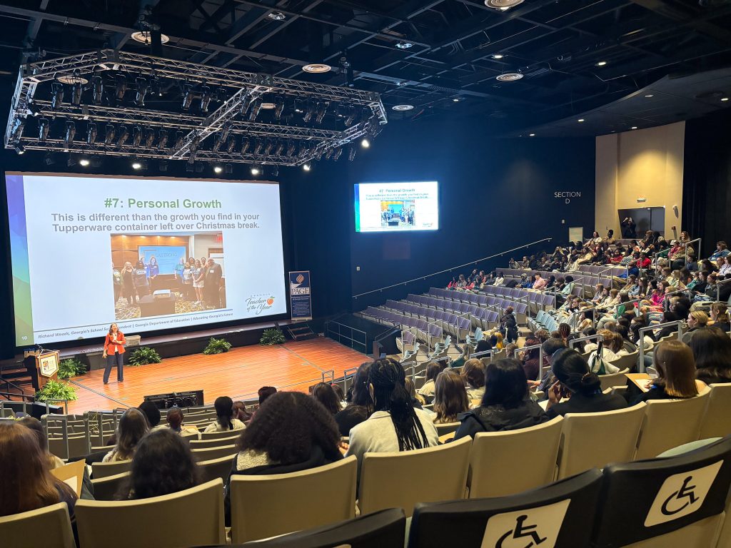 High school students sit in a large auditorium listening to a speaker on stage.