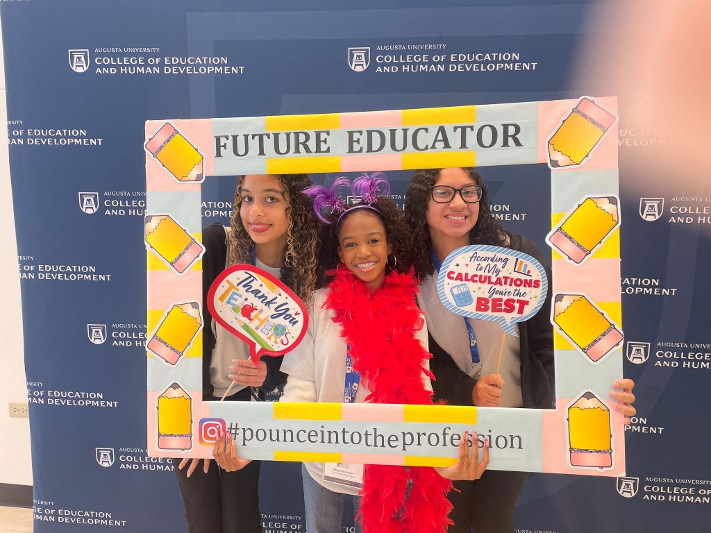 Three female high school students pose in a photo booth with different props, including an oversized picture frame with cutouts of pencils around the border and the words "Future Educator" at the top.