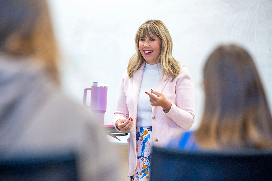 A female college professor stands at the front of a large college classroom an delivers a lecture to a group of students.