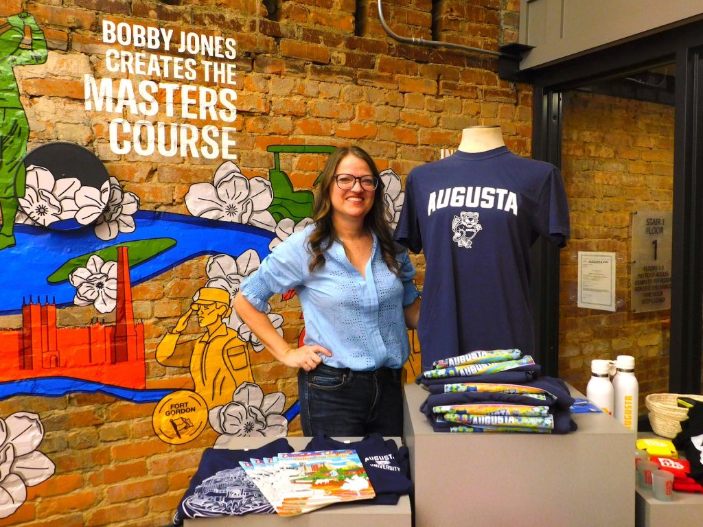 Woman standing in a store next to a shirt on display. Behind her is a mural on an old brick wall.