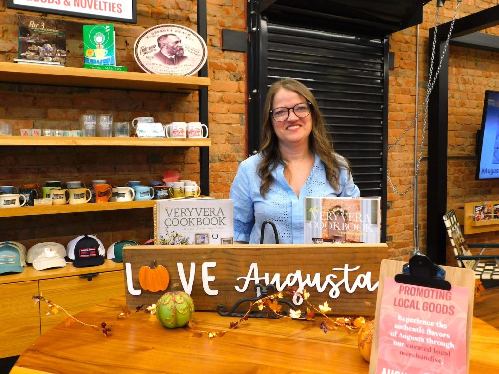 A woman who owns a small business stands inside her business with a sig that says, "Love Augusta." 