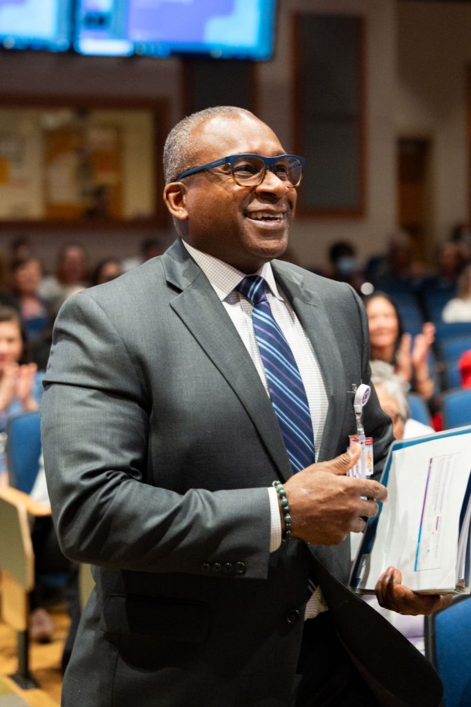 Man walking to a stage in a large, crowded auditorium. He is carrying a binder in his left hand.