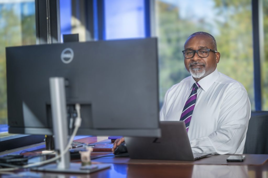 Man sitting at a desktop computer with a large window behind him.