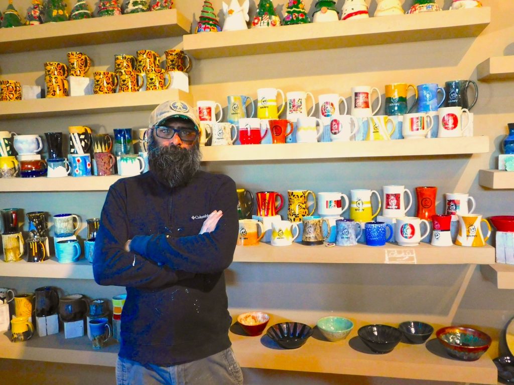 Man standing in a pottery shop with several shelves of ceramic mugs, cups and dishes behind him.