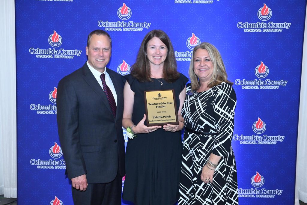 Two women and a man standing in front of a photo backdrop for Columbia County School District. The woman in the middle is holding a plaque for winning Teacher of the Year.