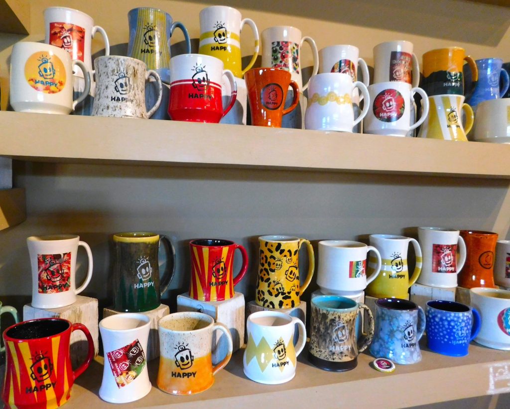 Two shelves filled with hand-made mugs, many have a smiling robot face and the word "Happy" on them.