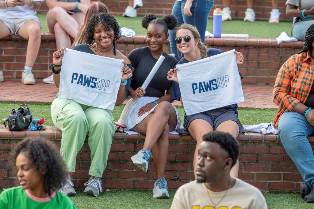 Three women sit on an outdoor ledge, and two of the women are holding up towels that read Paws Up.