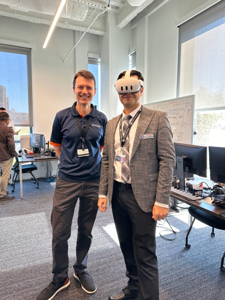 Two men stand in a large room. One is wearing an augmented reality headset.