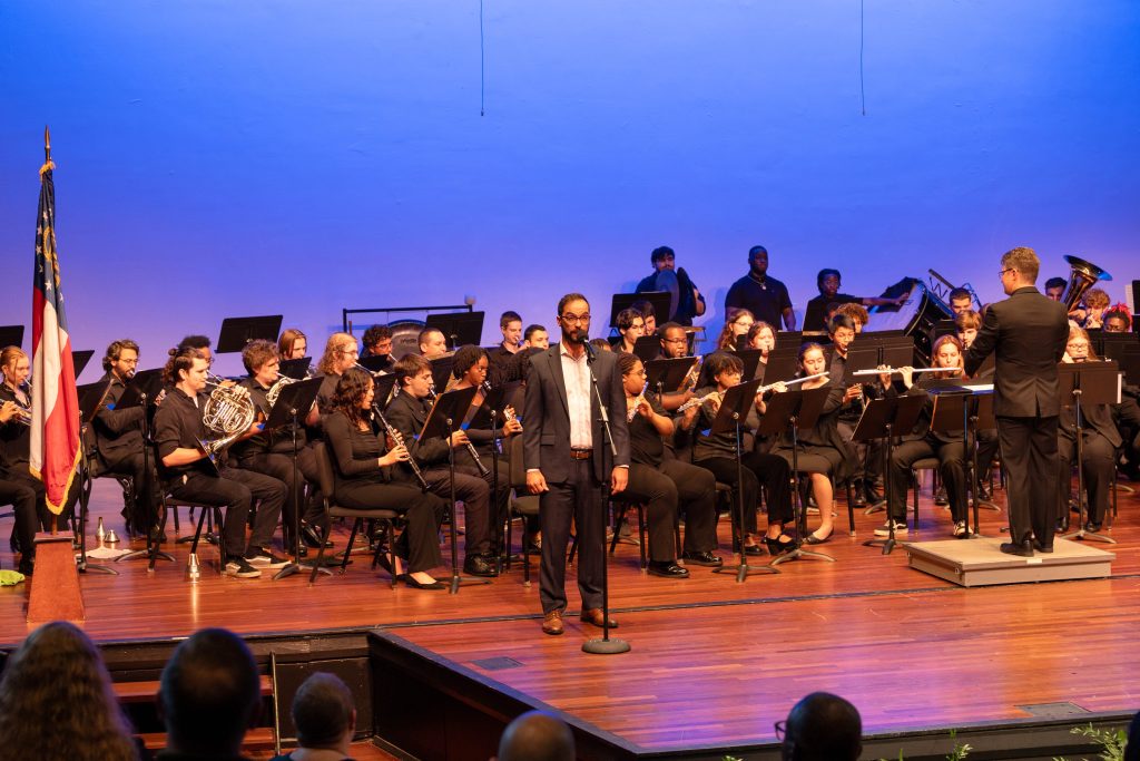 A man stands on stage in front of a microphone with a wind ensemble behind him.