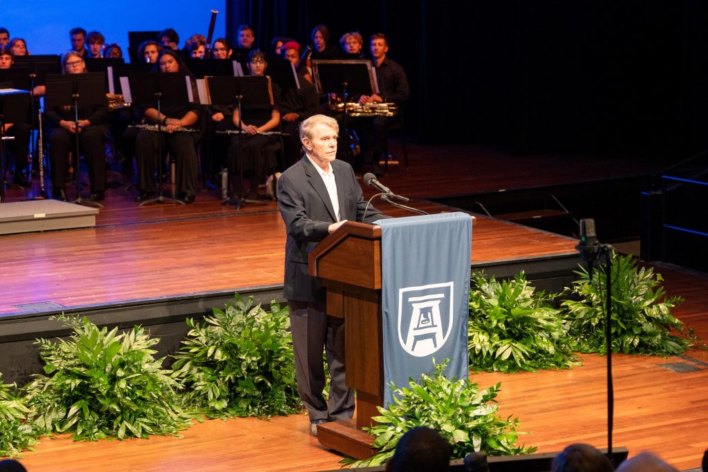 A man stands on stage behind a podium with a wind ensemble behind him.