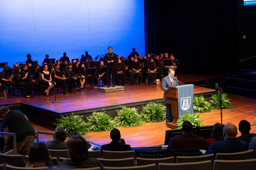 A man stands on stage behind a podium with a wind ensemble behind him.