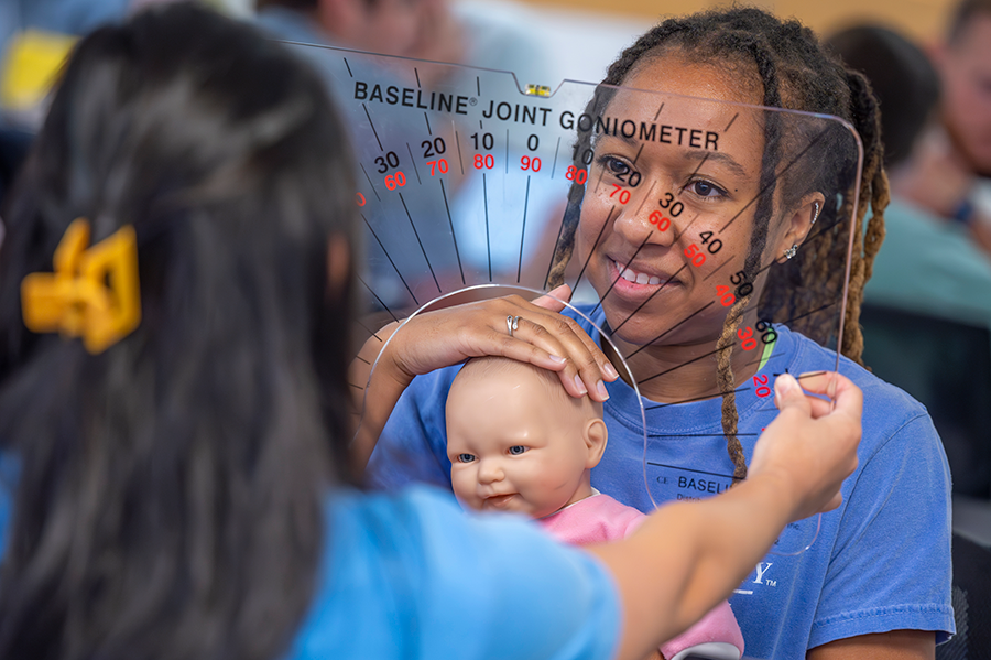 A female college student holds a mannequin of a baby and tilts its head to the side while another student holds up a chart to learn about joint movement.