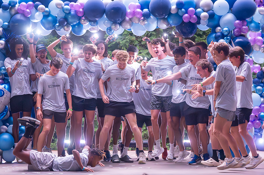 A large team of men's cross country runners cheer and film as a friend does the "worm" on the ground.