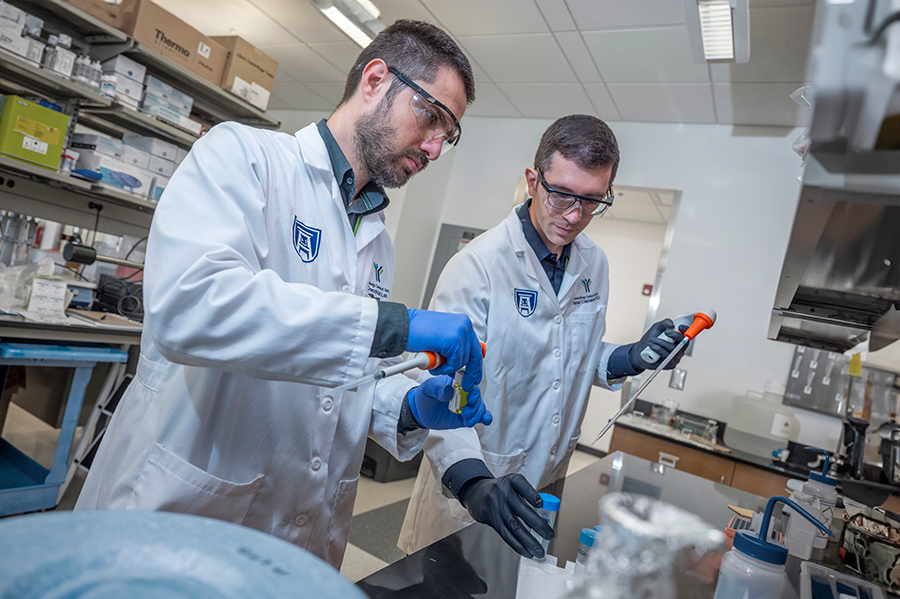 Two male researchers, both wearing safety glasses, gloves, and clinical coats, use test tubes to conduct experiments in a lab.