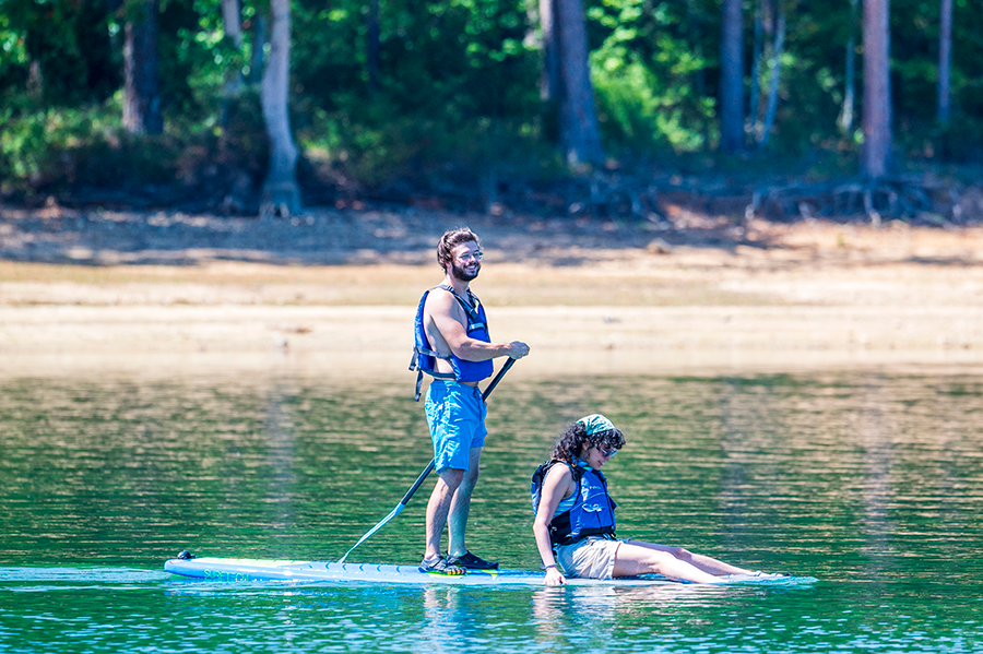 A college man and woman are on a paddle board in the middle of a lake. The man is standing and using the paddle to move them forward, while the woman is sitting with her legs in the water off the front of the board.