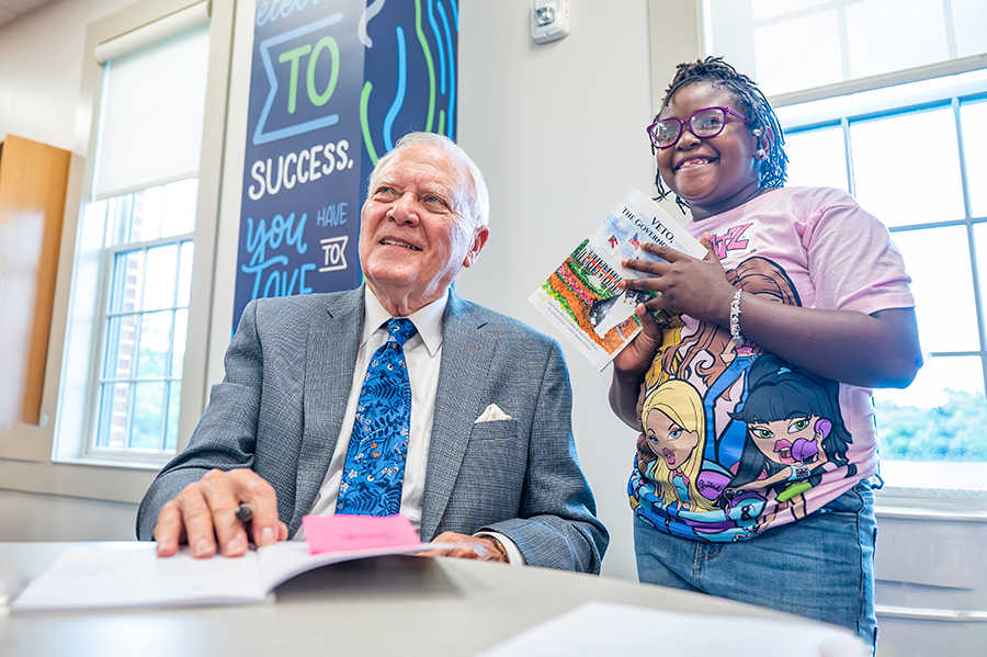 A man signs a book he wrote while a young child smiles and watches.