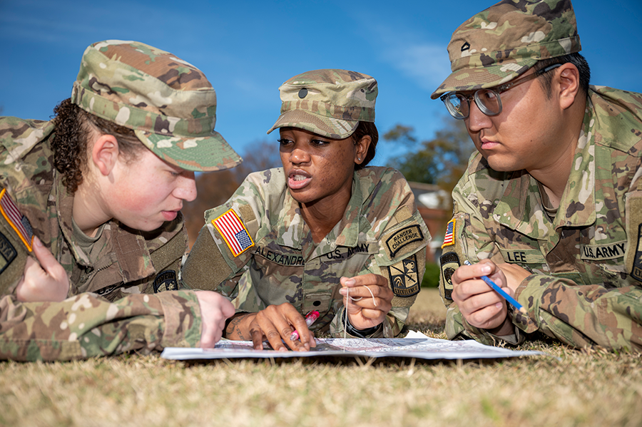 Three military members lay prone on the ground while looking at a map.