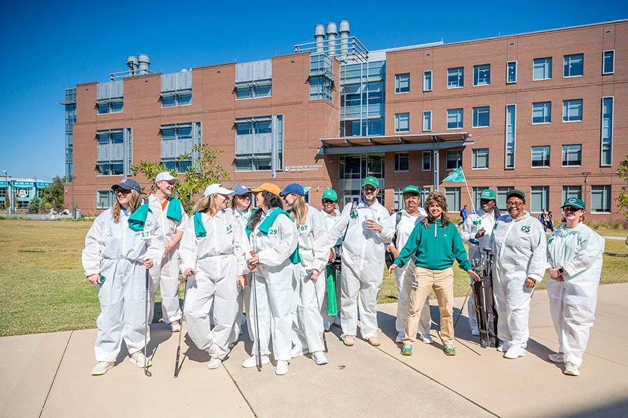 A large group of college students stand outside. They are all dressed like golf caddies with baseball caps and coveralls.