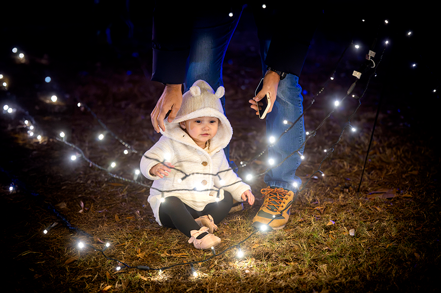 A toddler wearing a winter coat with fuzzy ears sits on the ground and looks at bright Christmas lights.