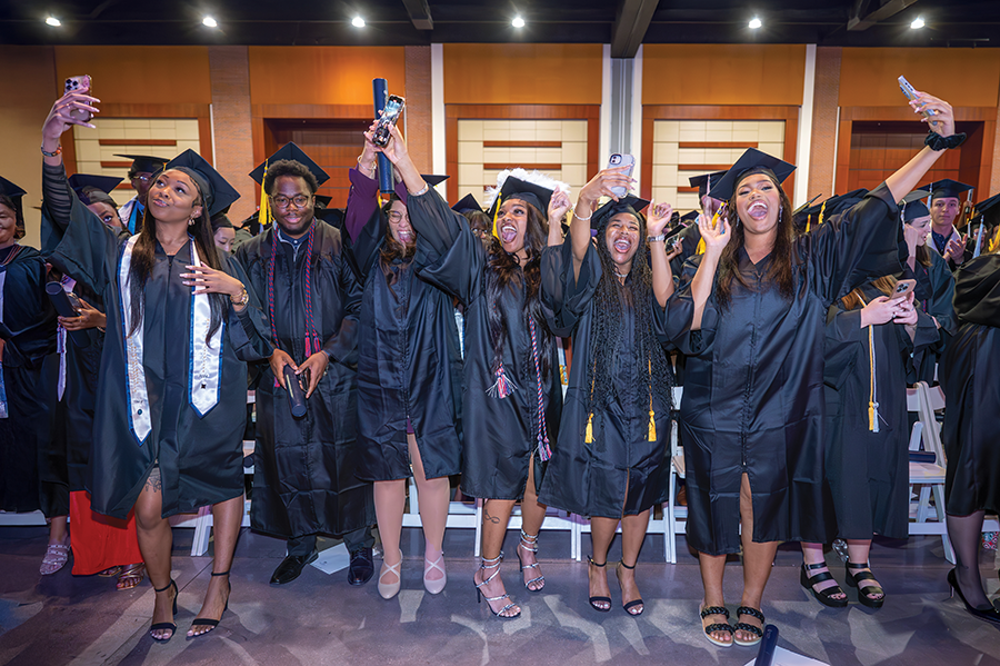 College graduates stand up and cheer, celebrating the completion of their graduation ceremony. Four are holding phones to film themselves.