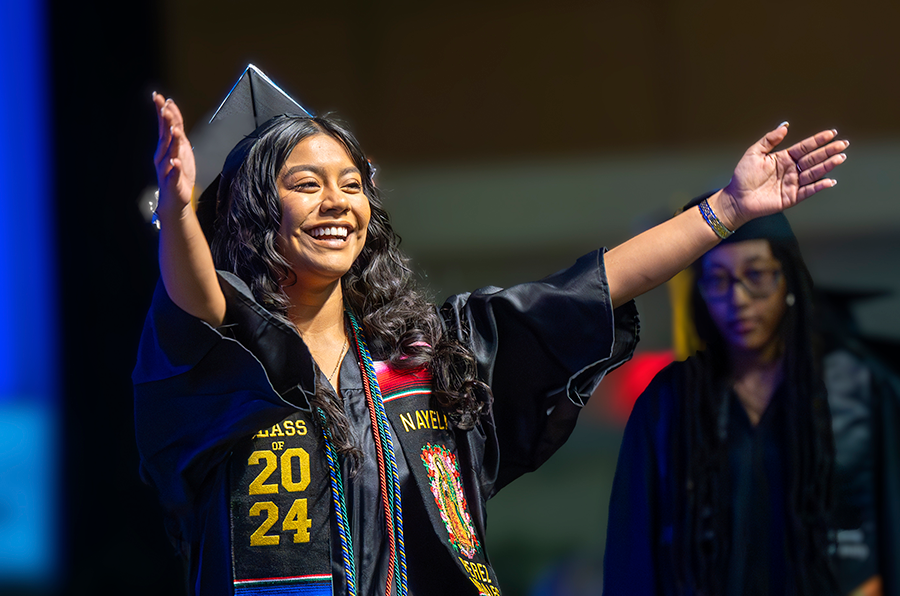 A college graduate in full graduation regalia holds her arms out for a hug as she walks across the graduation stage.