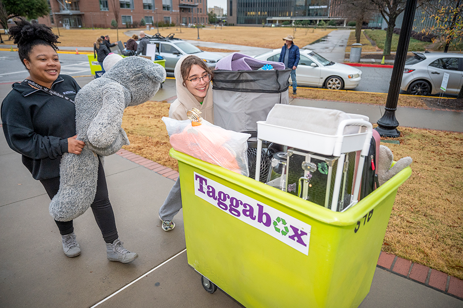 Two college women push a large box on wheels loaded with items for a college dorm.