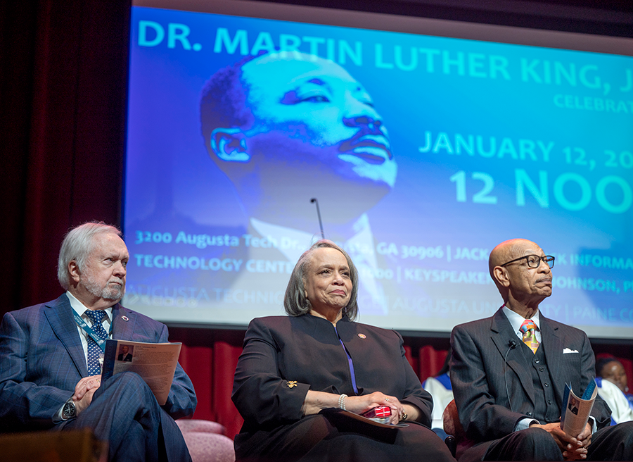 Three people sit on a stage with a large image of Martin Luther King Jr. on a projection screen behind them.