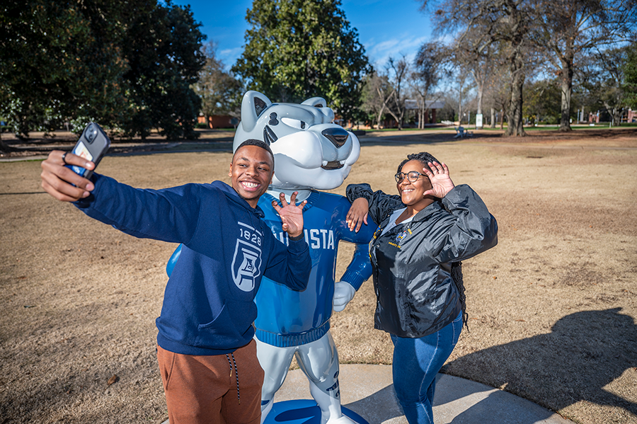 Two college students pose for a selfie with a large statue of a college mascot depicting a jaguar wearing a sweater.