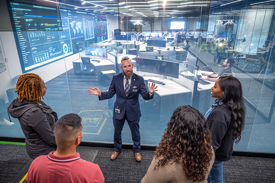 A man stands in front of a large window overlooking a big command center. The man is talking to three women and a man about what goes on in the command center.