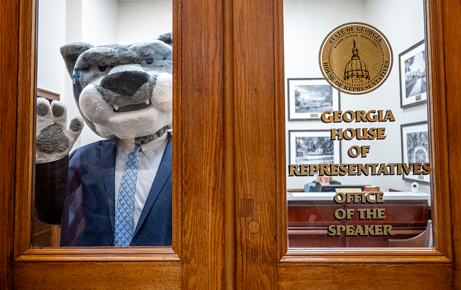 A college mascot depicting a jaguar wears a suit while standing behind a door with text on a window. The text reads, "Georgia House of Representatives, Office of the Speaker."