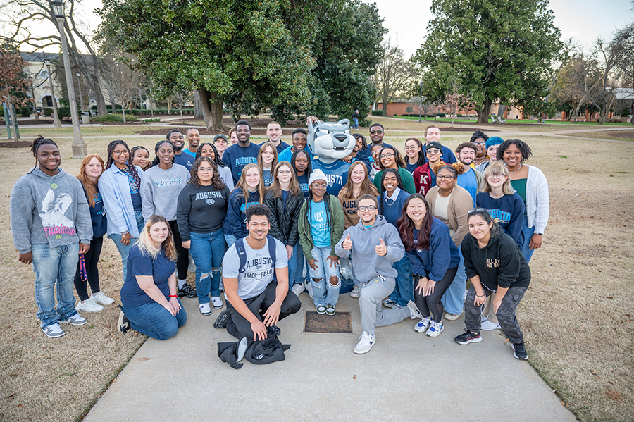 A large group of college students gather around a statue depicting a college mascot in the shape of a Jaguar. The statue is wearing a sweater with the word "Augusta" across the chest.