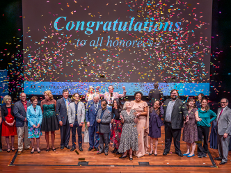 A large group of college professors stand on a stage as confetti falls from above them.