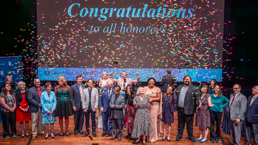 A large group of college professors stand on a stage as confetti falls from above them.