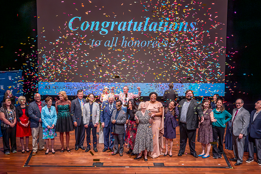 A large group of college professors stand on a stage as confetti falls from above them.