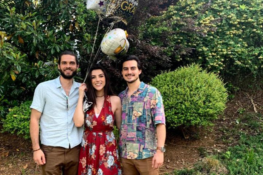 Two men and a woman, all siblings, stand outside. The woman is holding strings attached to four balloons that are floating above her head.