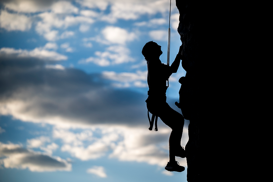 A male college student climbs an outdoor climbing wall while attached to a harness.