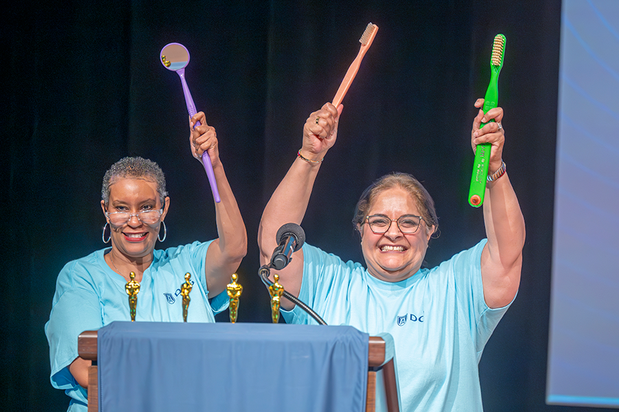 Two college professors stand on a stage and hold oversized toothbrushes.