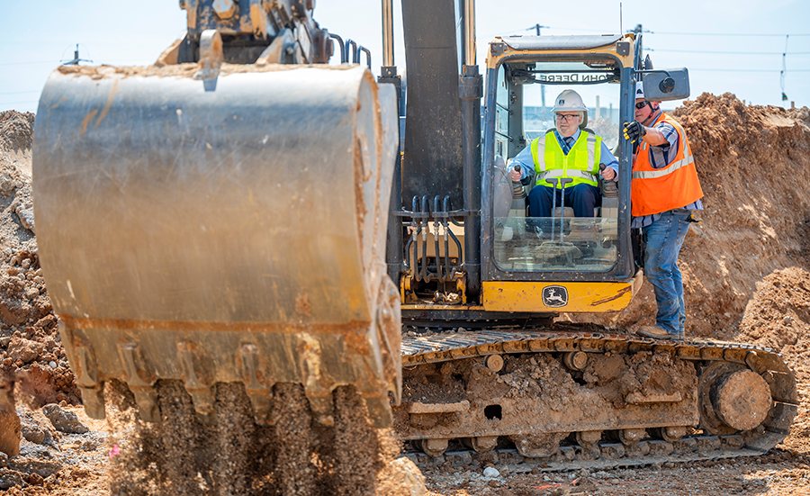 A man in a safety vest and hard hat sits at the controls of a large piece of equipment. Another man is standing to his side instructing him how to operate the machine.