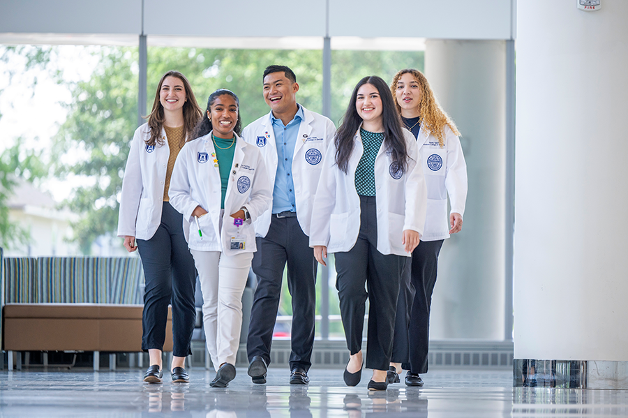 Five medical students wearing their clinical coats walk through a hallway while talking.